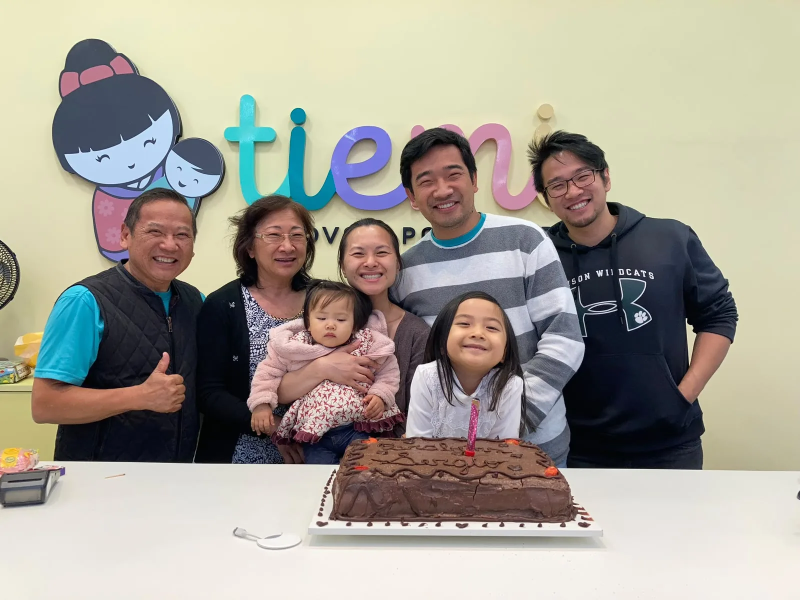 Família Tiemi reunida em comemoração na loja, com três gerações posando em frente à logo da Tiemi